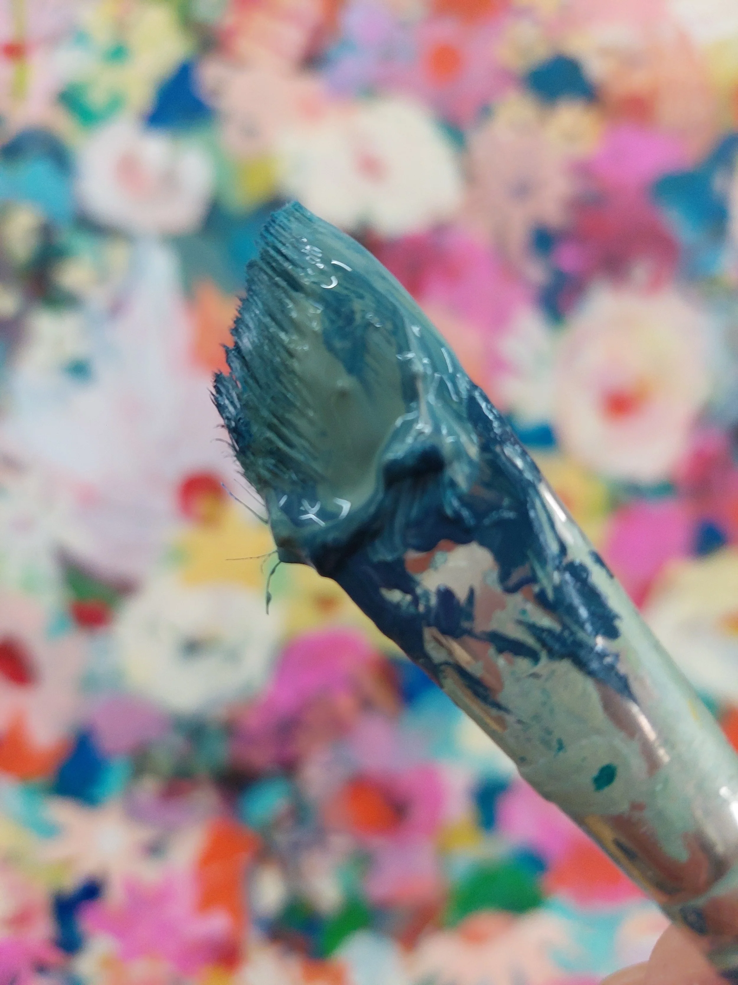 Close-up of a paintbrush covered in blue and green paint against a colorful, abstract background of soft pastel colors and out-of-focus floral shapes.