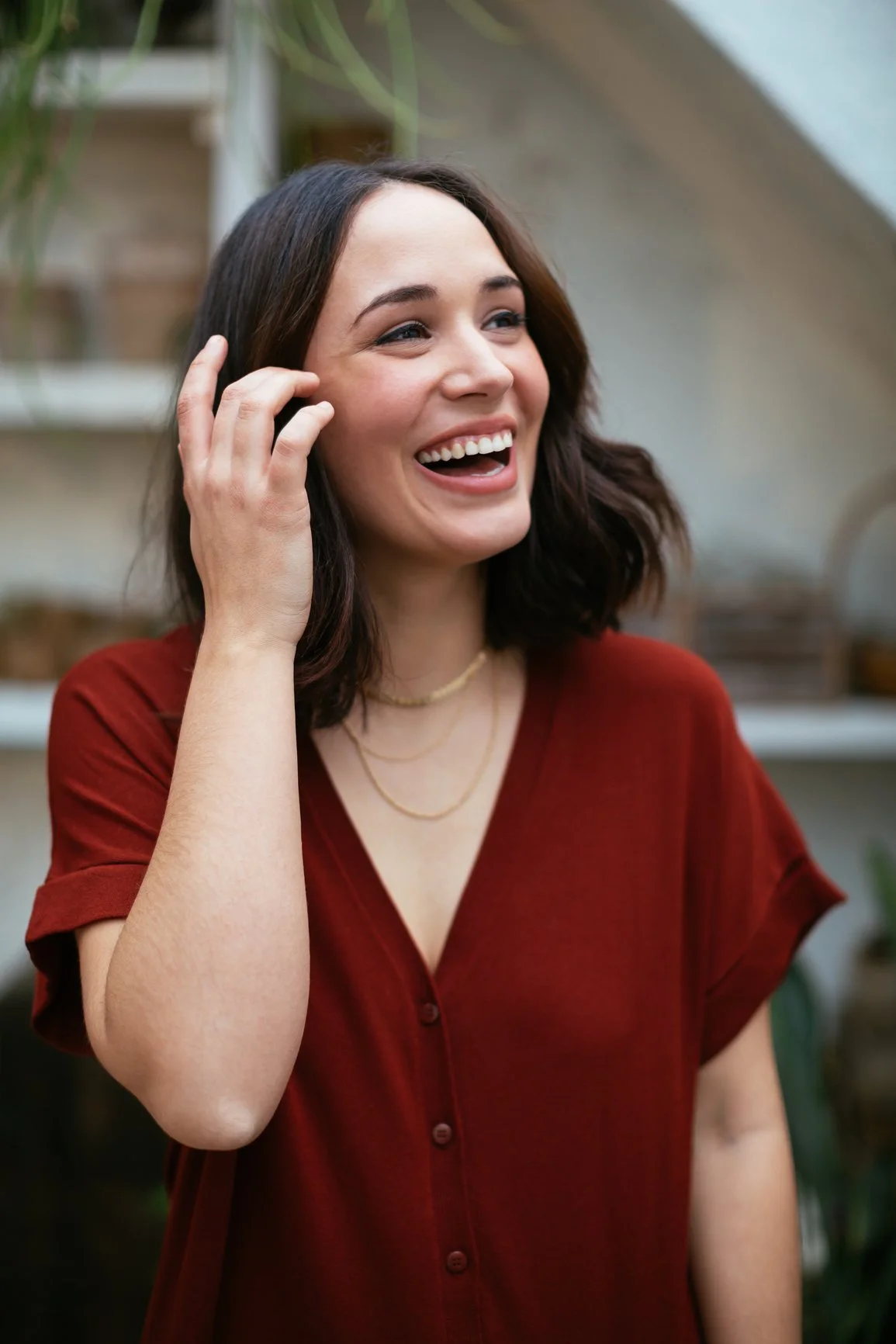 A woman with dark brown hair, wearing a red shirt and gold necklaces, smiling and touching her face in a cheerful moment.