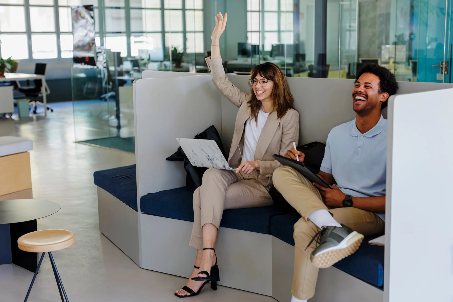 Two colleagues sitting in a semi-private booth in an office, one woman raising her hand and smiling, the man sitting next to her also smiling, holding a tablet and a pen.
