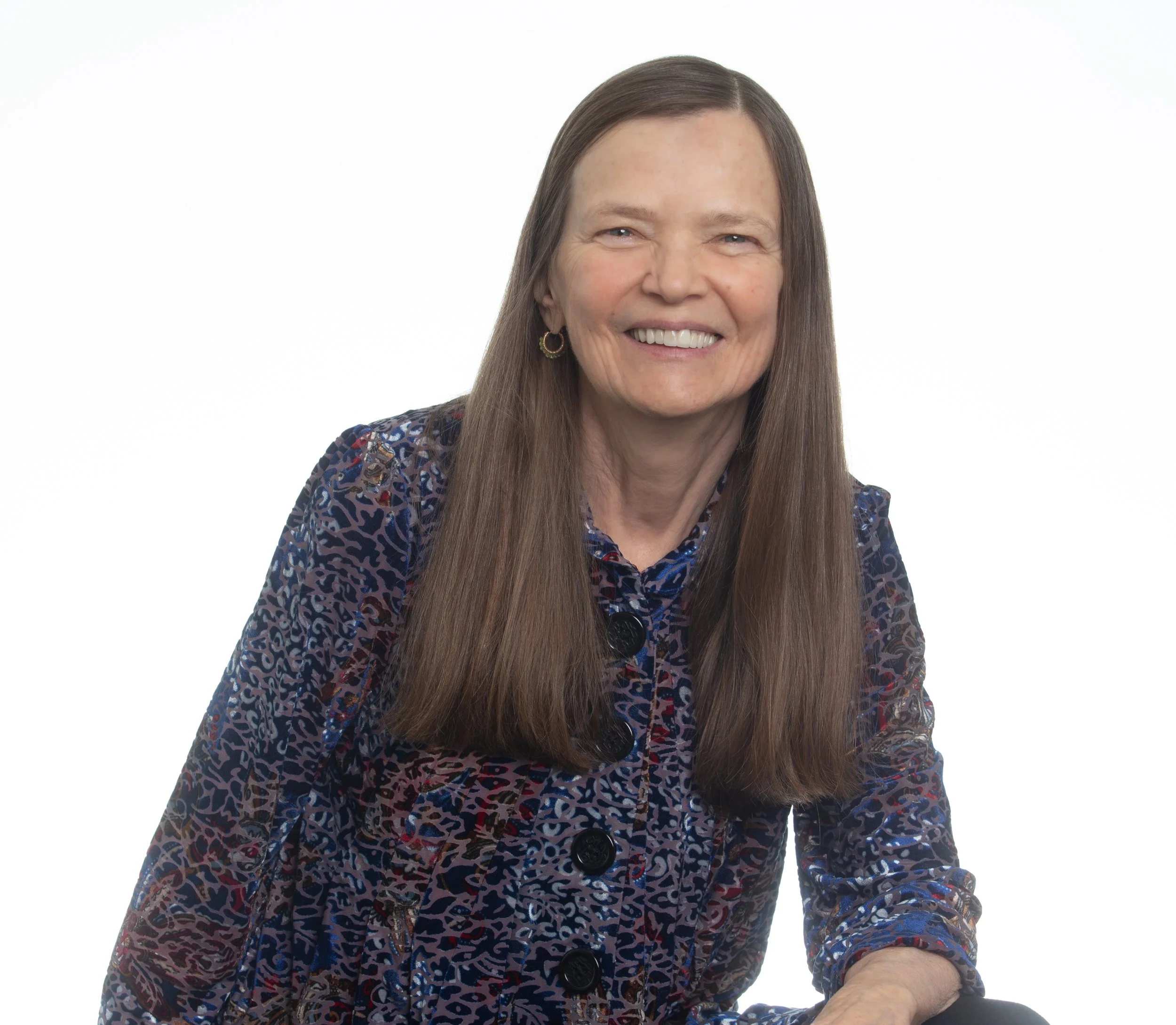 Portrait of a woman with long brown hair, smiling, wearing a patterned blouse and gold earrings, against a white background.