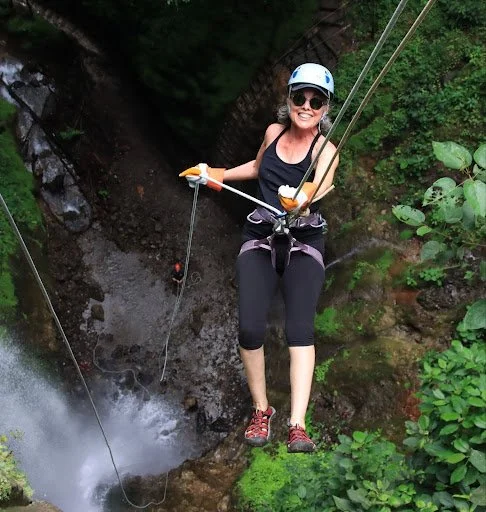 Woman in safety gear zip-lining over a waterfall in a forest.