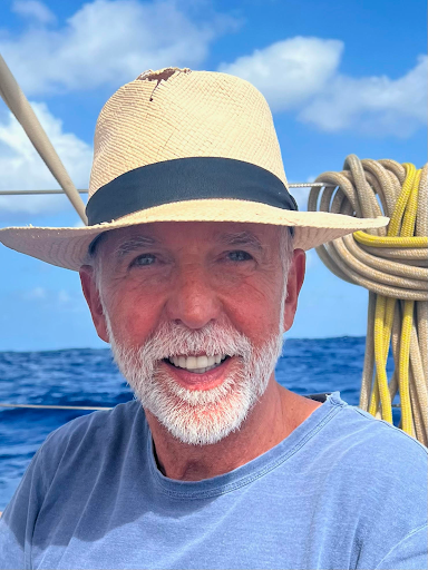 A smiling elderly man with a white beard and mustache wearing a straw hat and a blue shirt, on a boat with the sea and blue sky in the background.