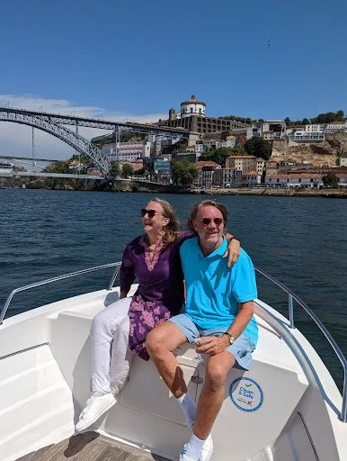 Two people sitting together on a boat, with a cityscape and bridge in the background, under a blue sky.