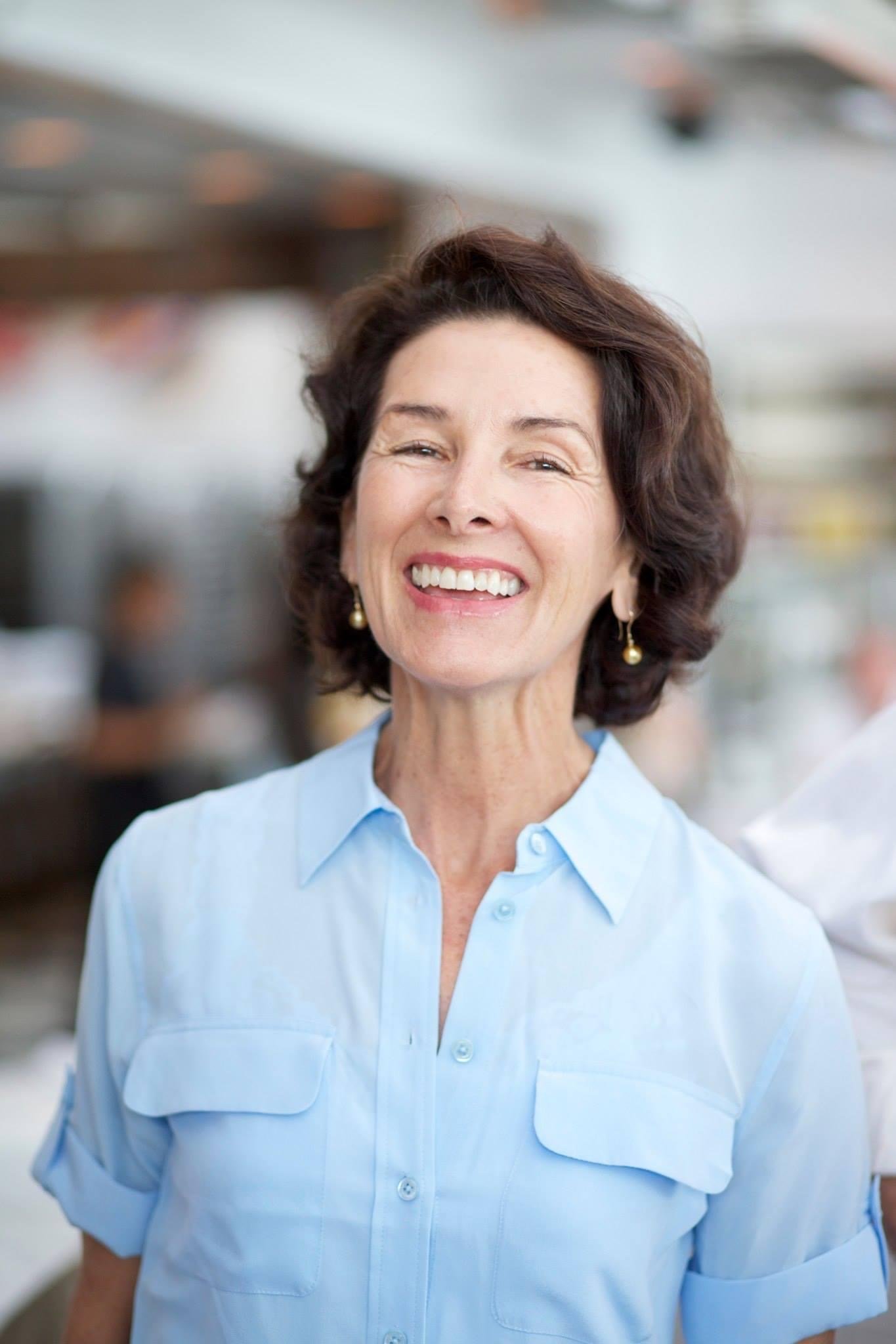 Smiling woman with short brown hair and earrings, wearing a light blue button-up shirt, in a bright indoor setting.