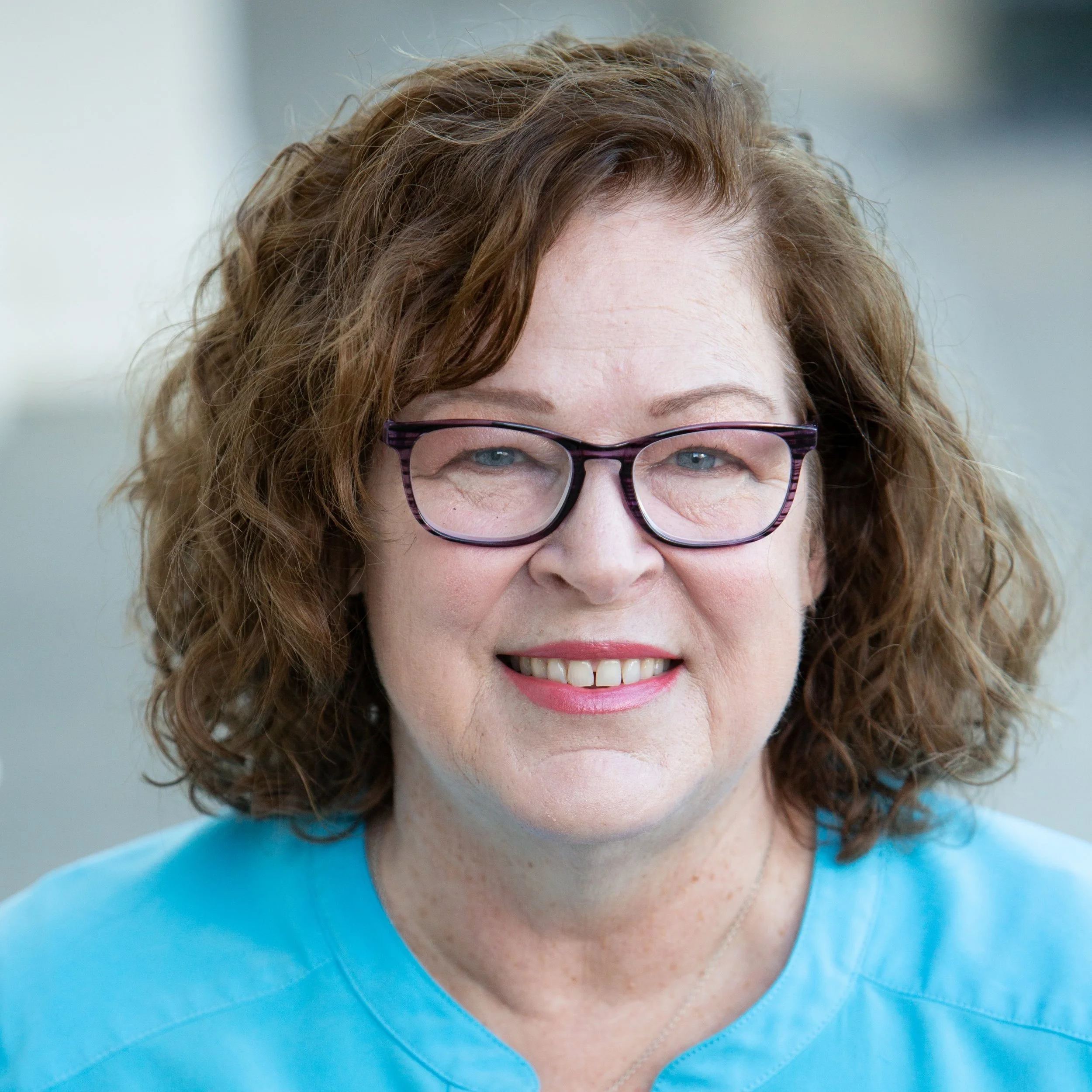 A smiling woman with curly red hair, wearing glasses and a blue top, outdoors during daytime.