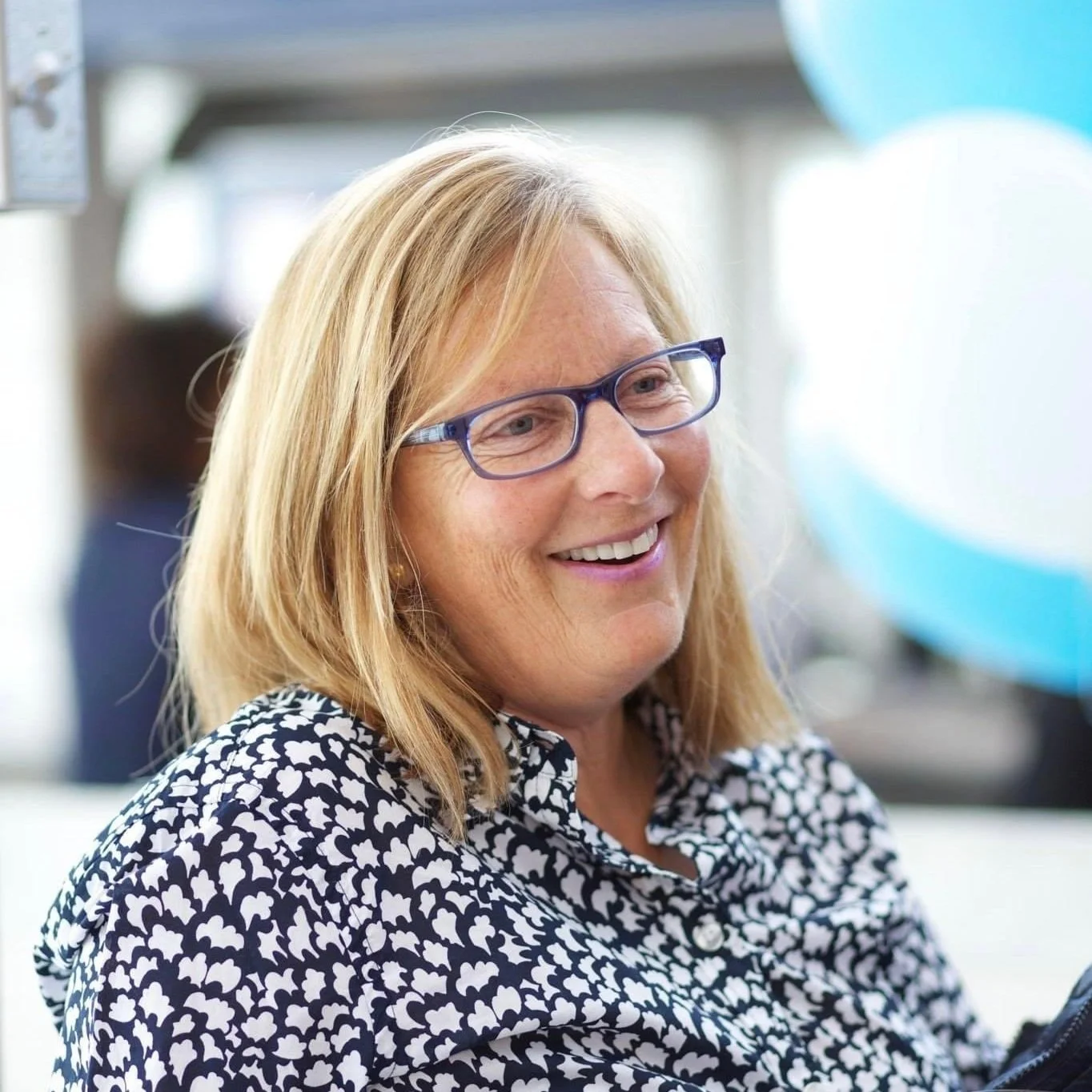 A woman with blonde hair and glasses smiling while looking slightly to her right, wearing a black and white patterned blouse, with a blurry background including balloons.