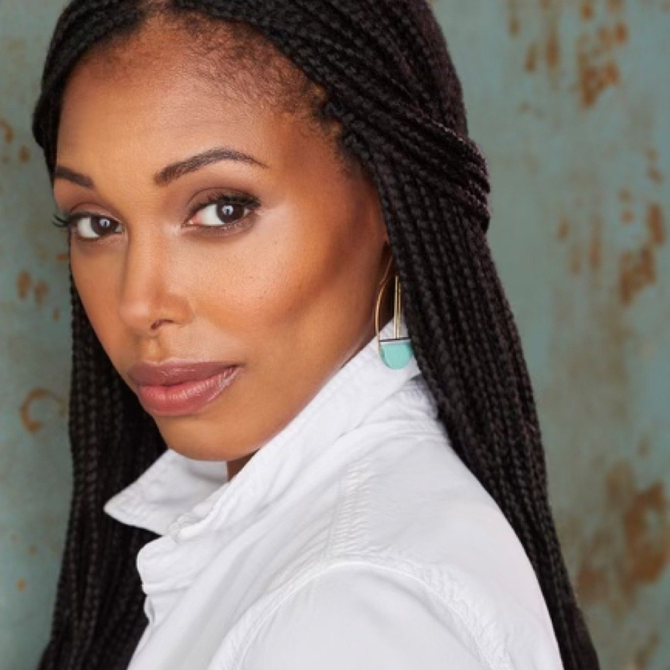 Close-up of an African American woman with braided hair, wearing a white collared shirt and turquoise and gold earrings, looking at the camera against a weathered, rusty background.