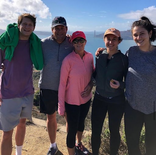 Group of five people standing outdoors near water with a bridge in the background.