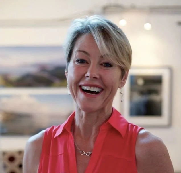 A woman with short gray hair smiling, wearing a sleeveless red blouse and jewelry, in an indoor setting.