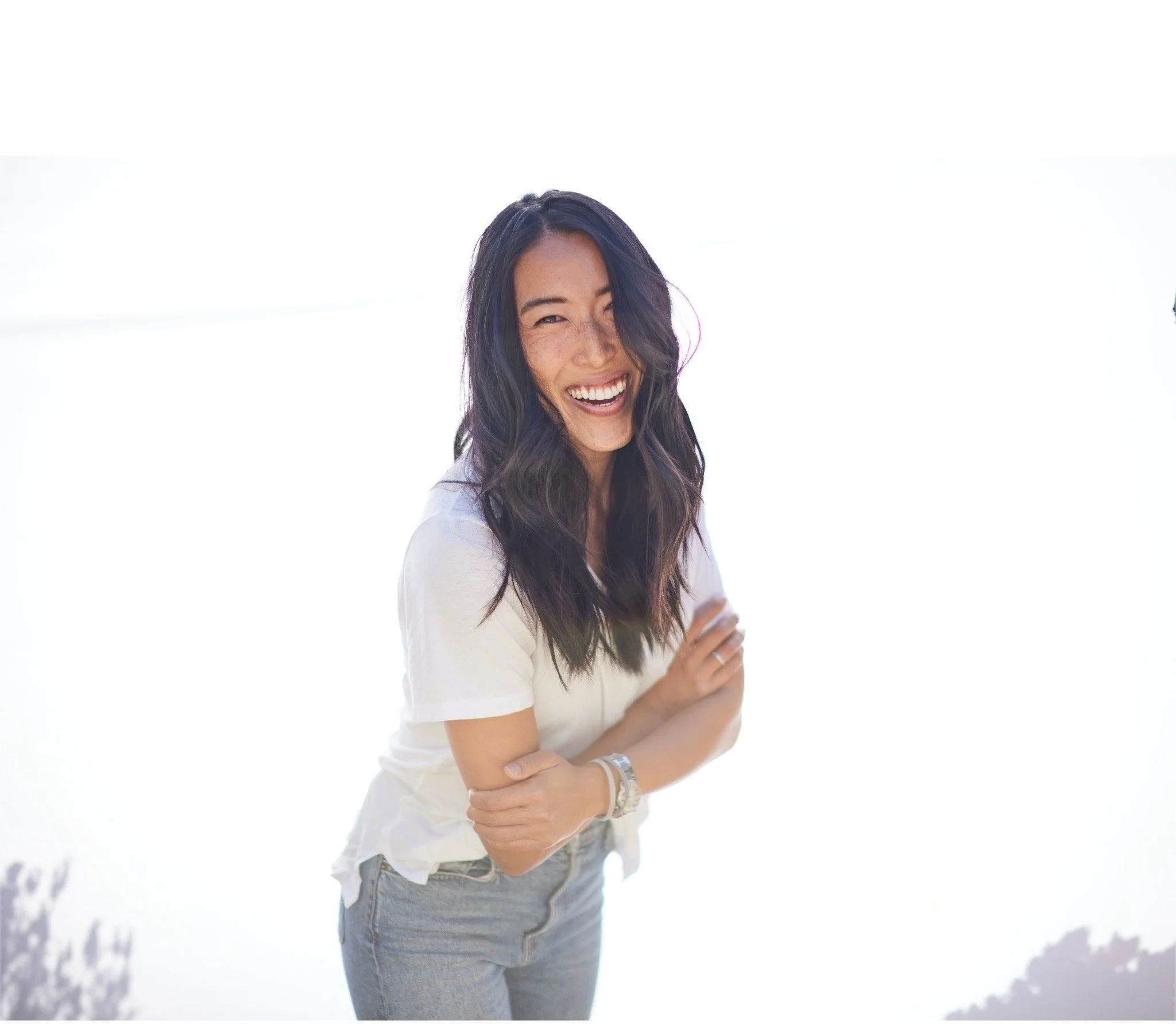 Smiling woman with dark wavy hair wearing a white t-shirt and gray jeans standing against a bright background.
