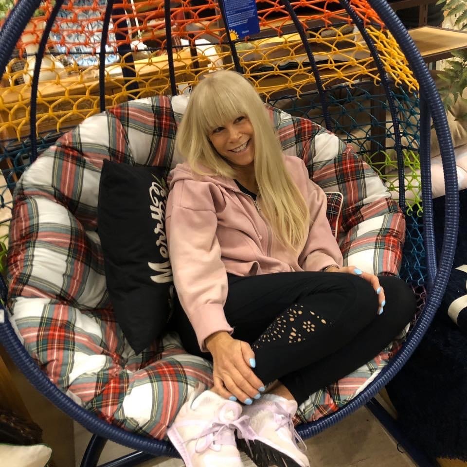 A woman with long blonde hair sitting in a circular hanging chair with a plaid cushion, smiling in a store with various colorful home decor items around her.