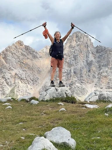 Person standing on a rock with arms raised holding trekking poles, smiling, in front of mountain cliffs.
