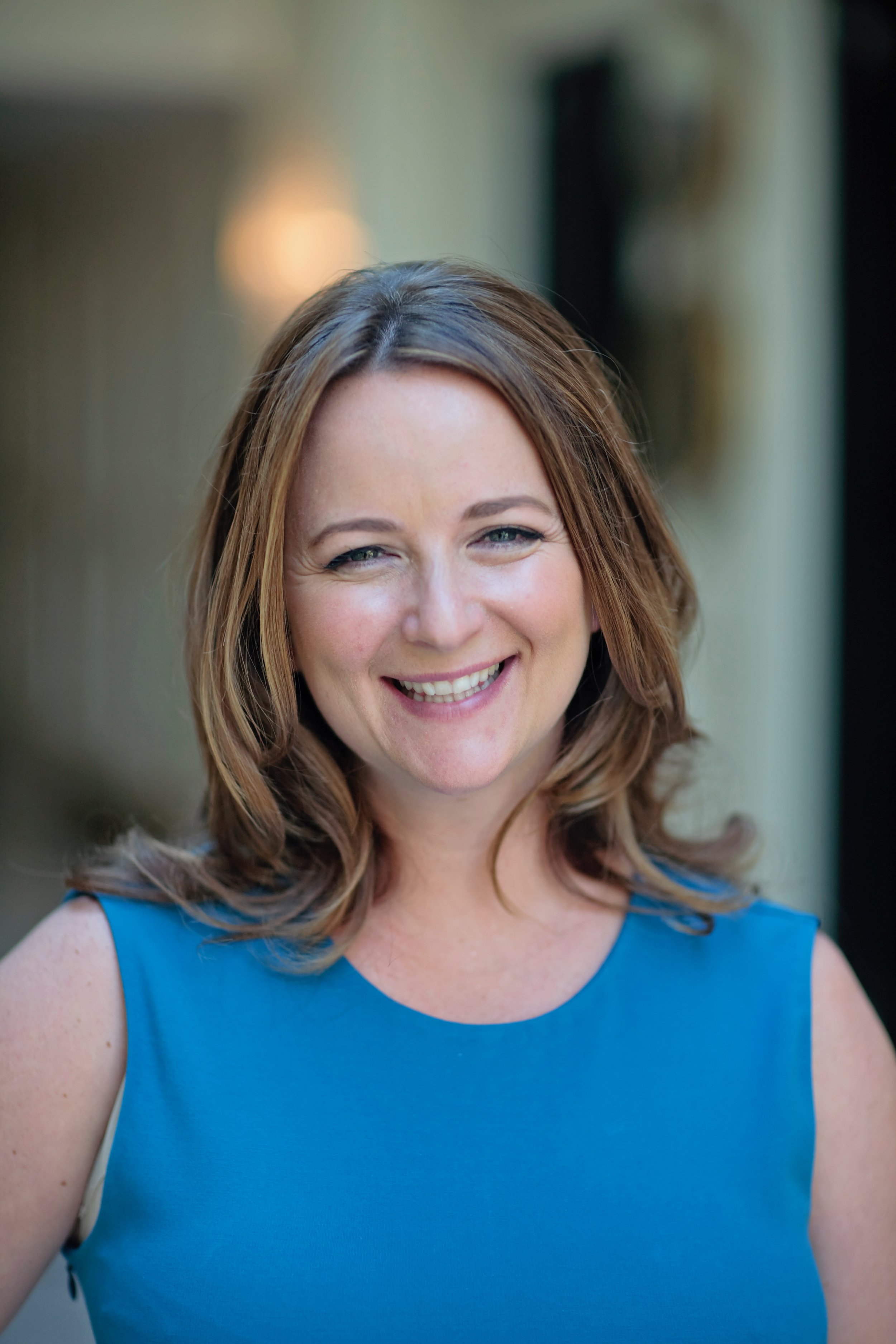 A smiling woman with shoulder-length light brown hair, wearing a sleeveless blue top, standing outdoors with a blurred background.