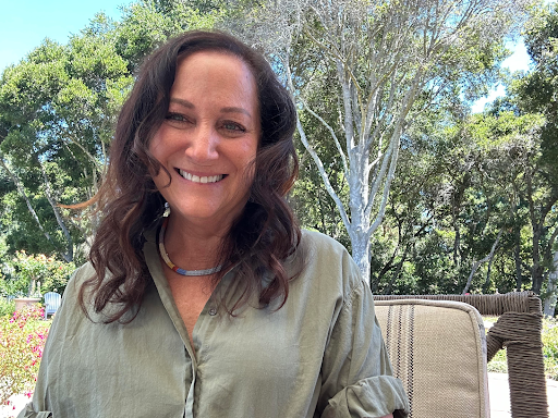 A woman with long wavy brown hair smiling outdoors, wearing a light green shirt, with trees and a wicker chair in the background.