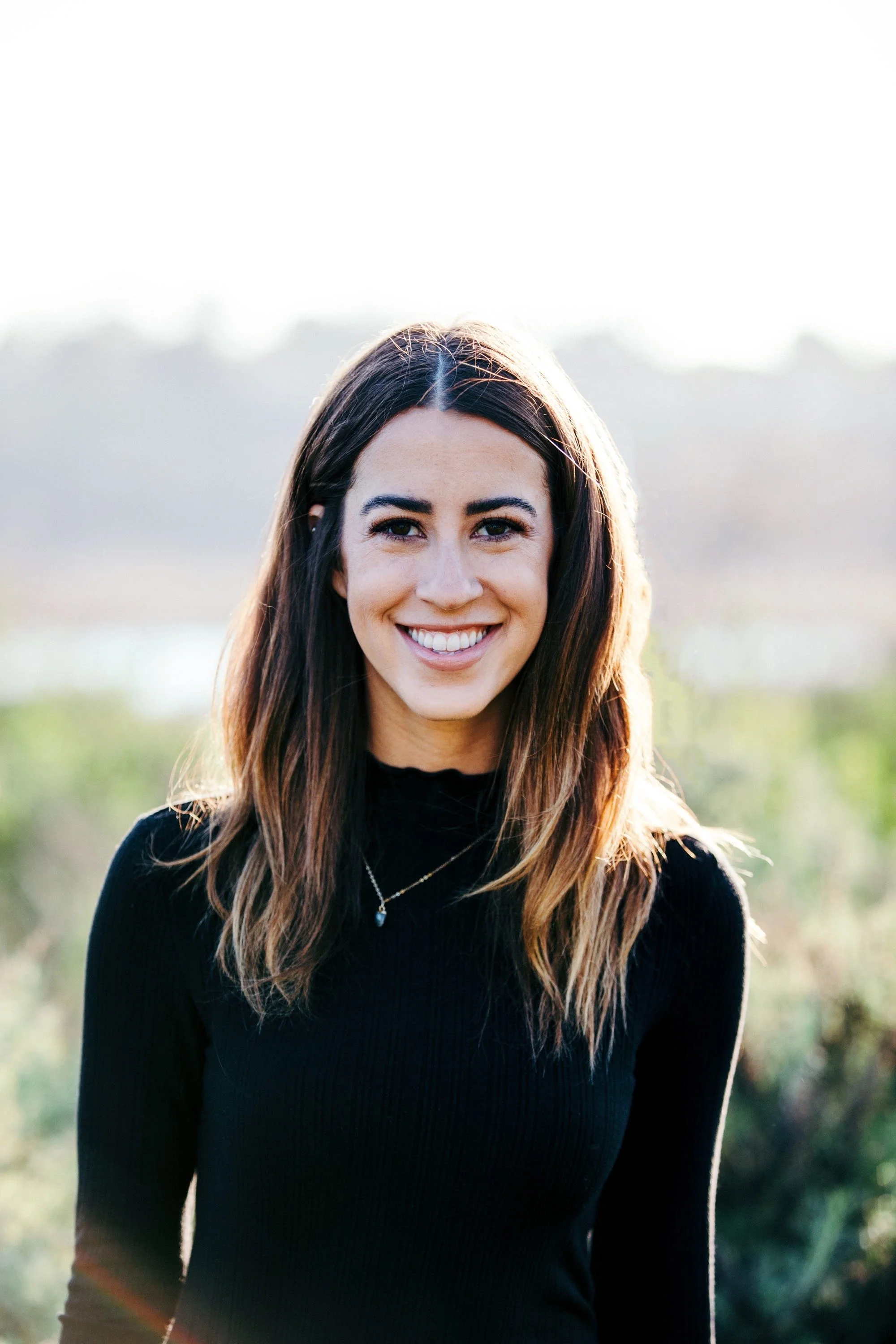 A woman with long brown hair smiling outdoors in front of a blurred natural background.