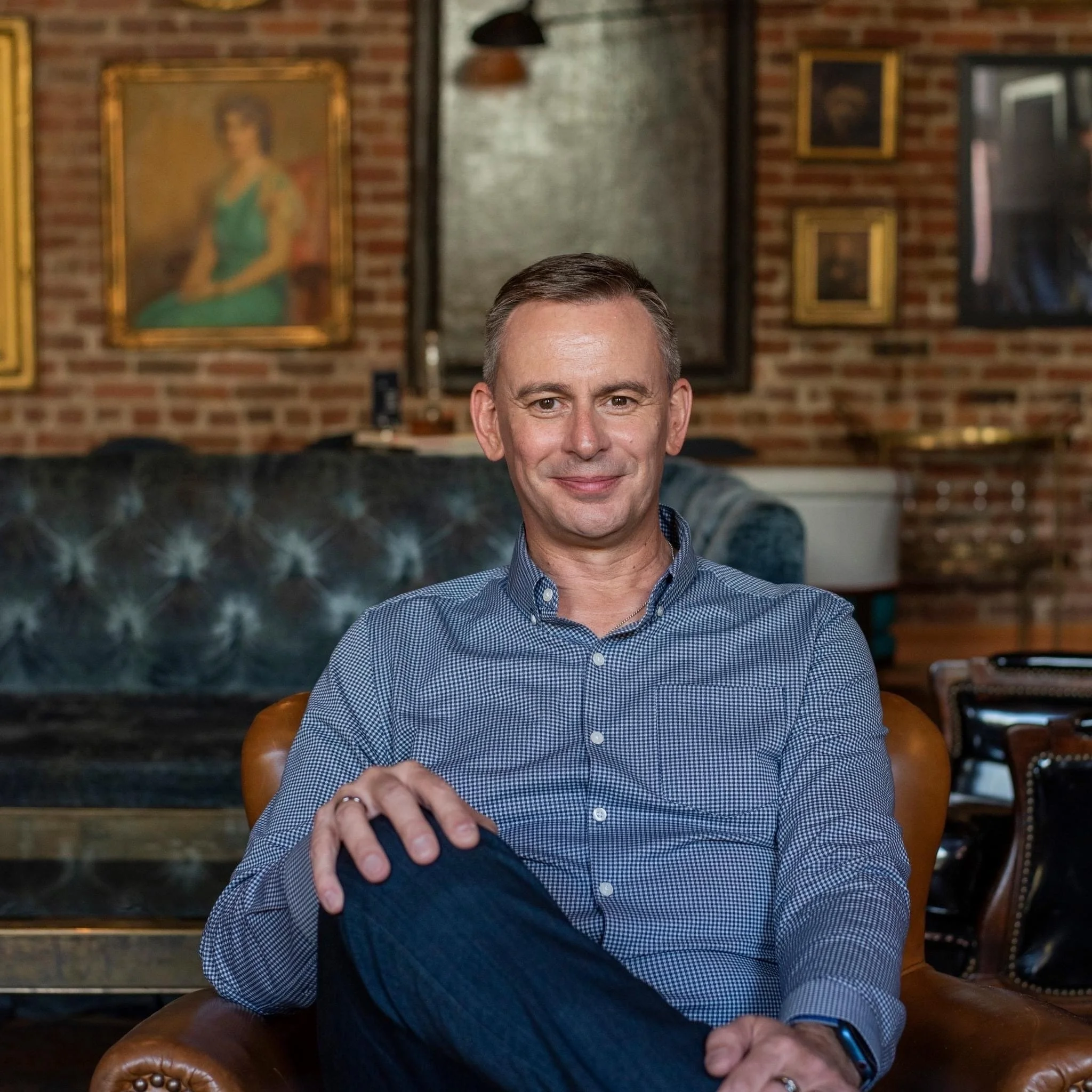 A middle-aged man with short brown hair, wearing a blue checkered shirt, sitting in a brown leather chair in a cozy, art-filled room with brick walls.