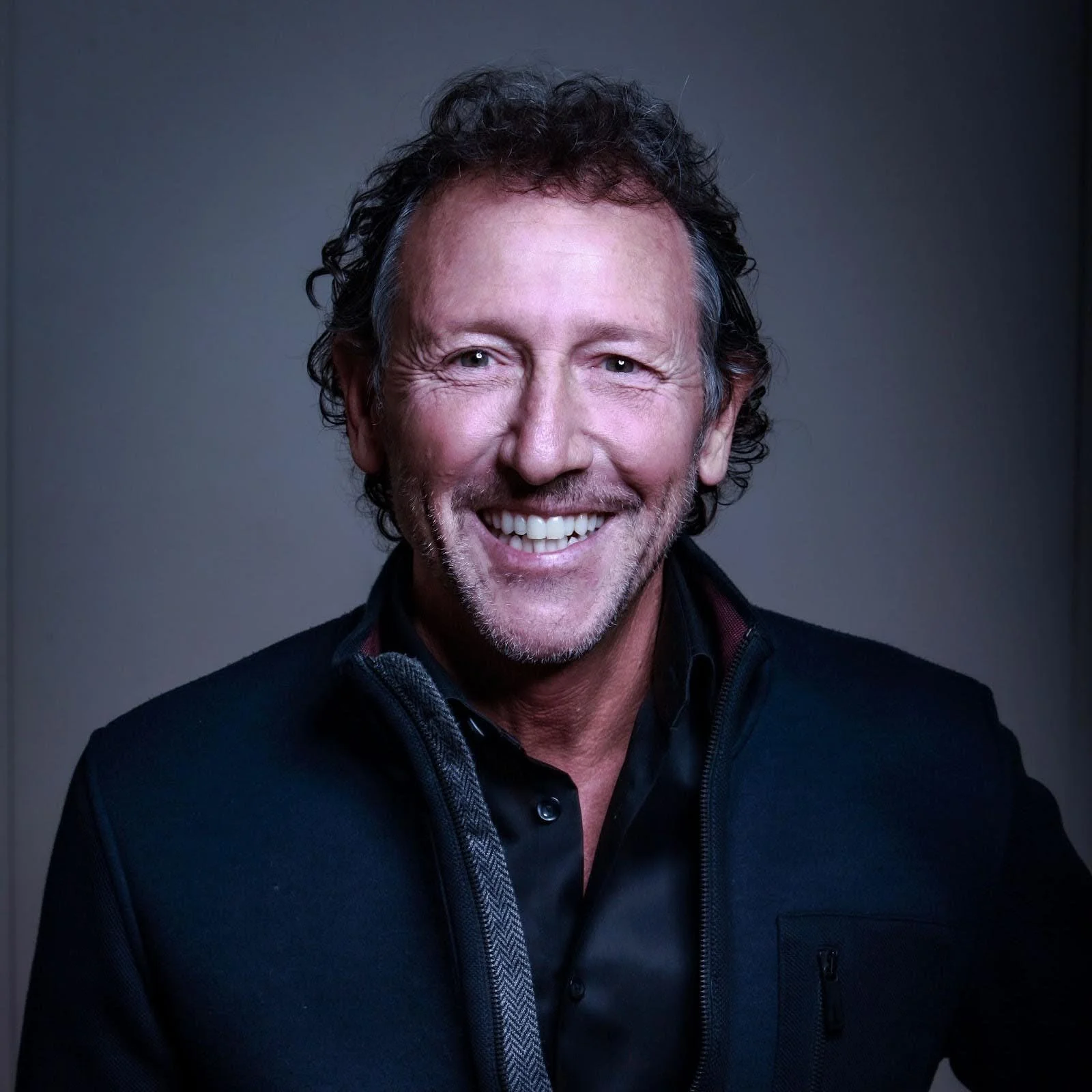 Close-up of smiling middle-aged man with curly hair, wearing a black jacket and dark shirt, against a dark background.