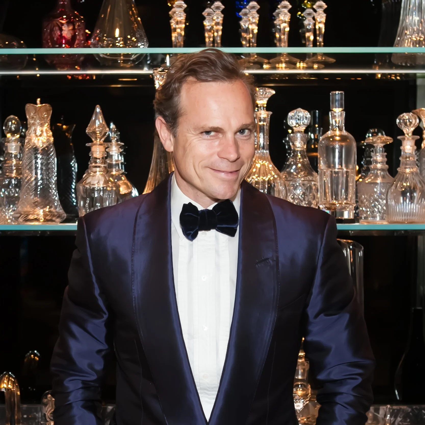 A man in a tuxedo with a black bow tie, smiling, standing in front of a display of glass decanters and bottles.