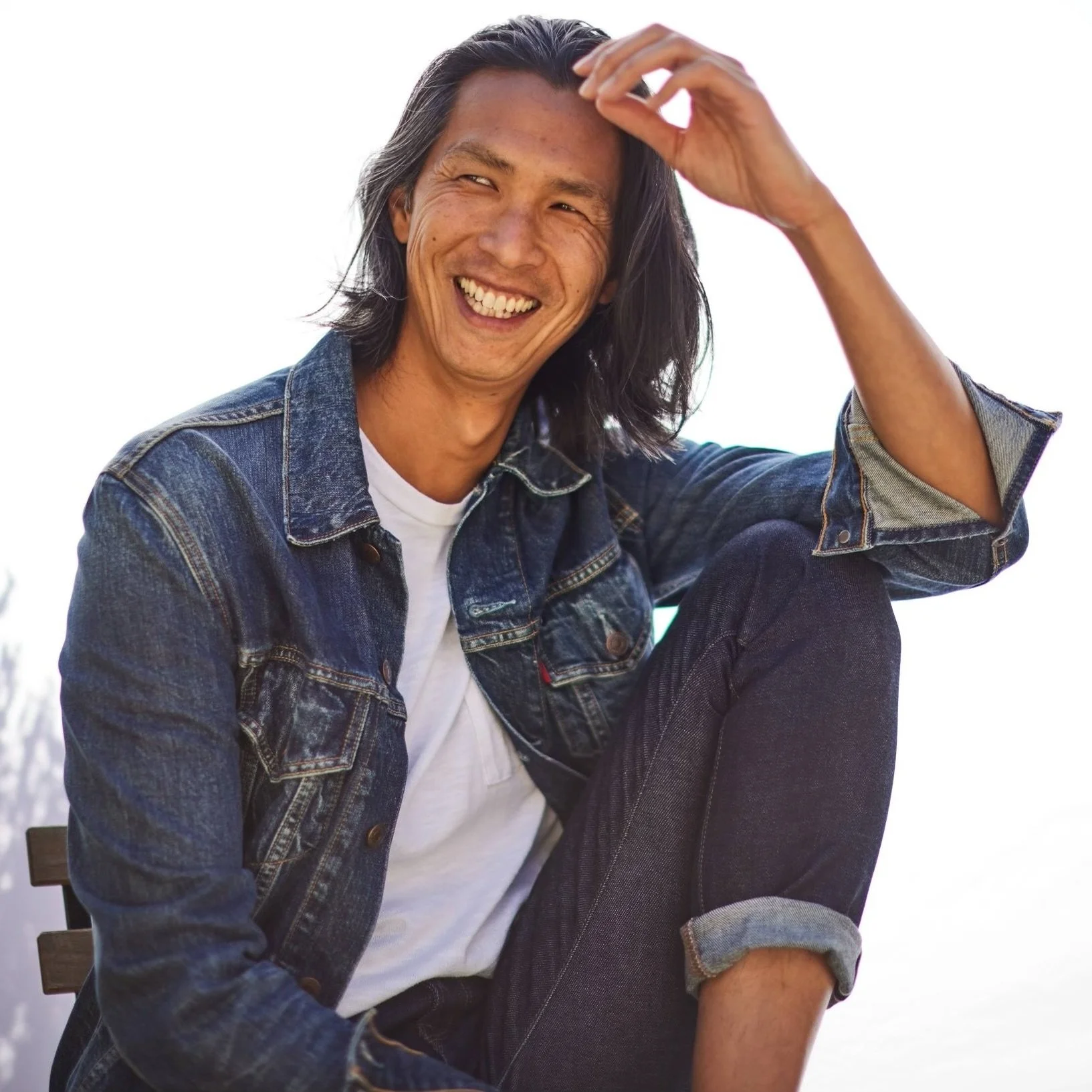 A smiling man with shoulder-length black hair, wearing a blue denim jacket over a white T-shirt, sitting outdoors, with a bright sky in the background.