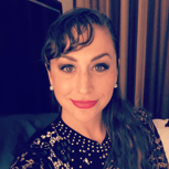 A woman with dark, wavy hair and bold makeup, wearing a patterned top, smiling at the camera indoors.