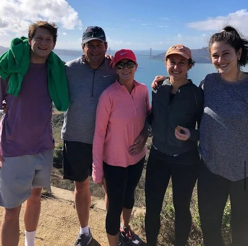 Group of five people standing outdoors near water with a bridge in the background.