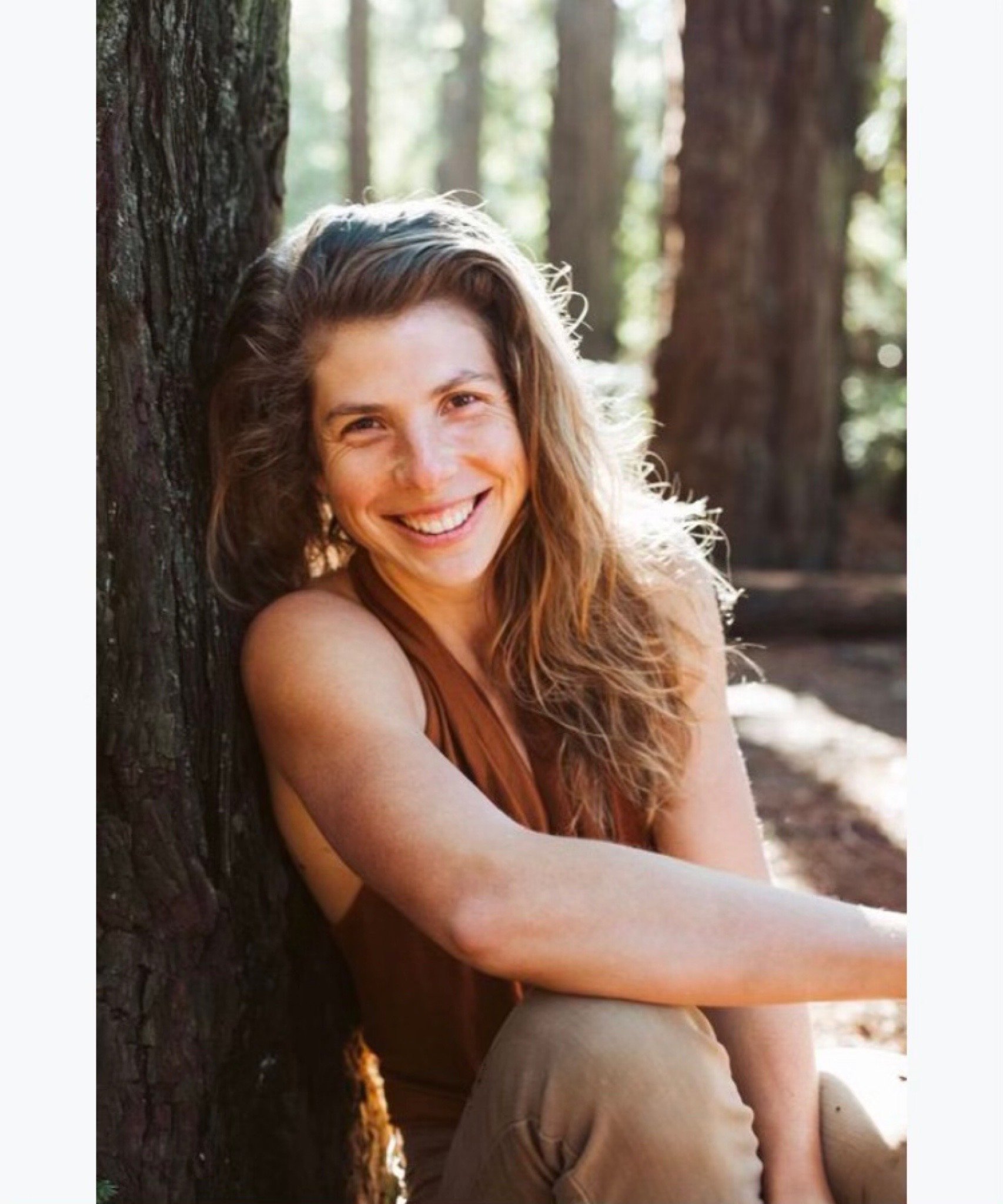 Smiling woman sitting outdoors leaning against a tree in a forest