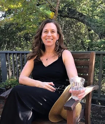 Woman sitting on a wooden outdoor chair, holding a glass of rose water, smiling, with green trees in the background.