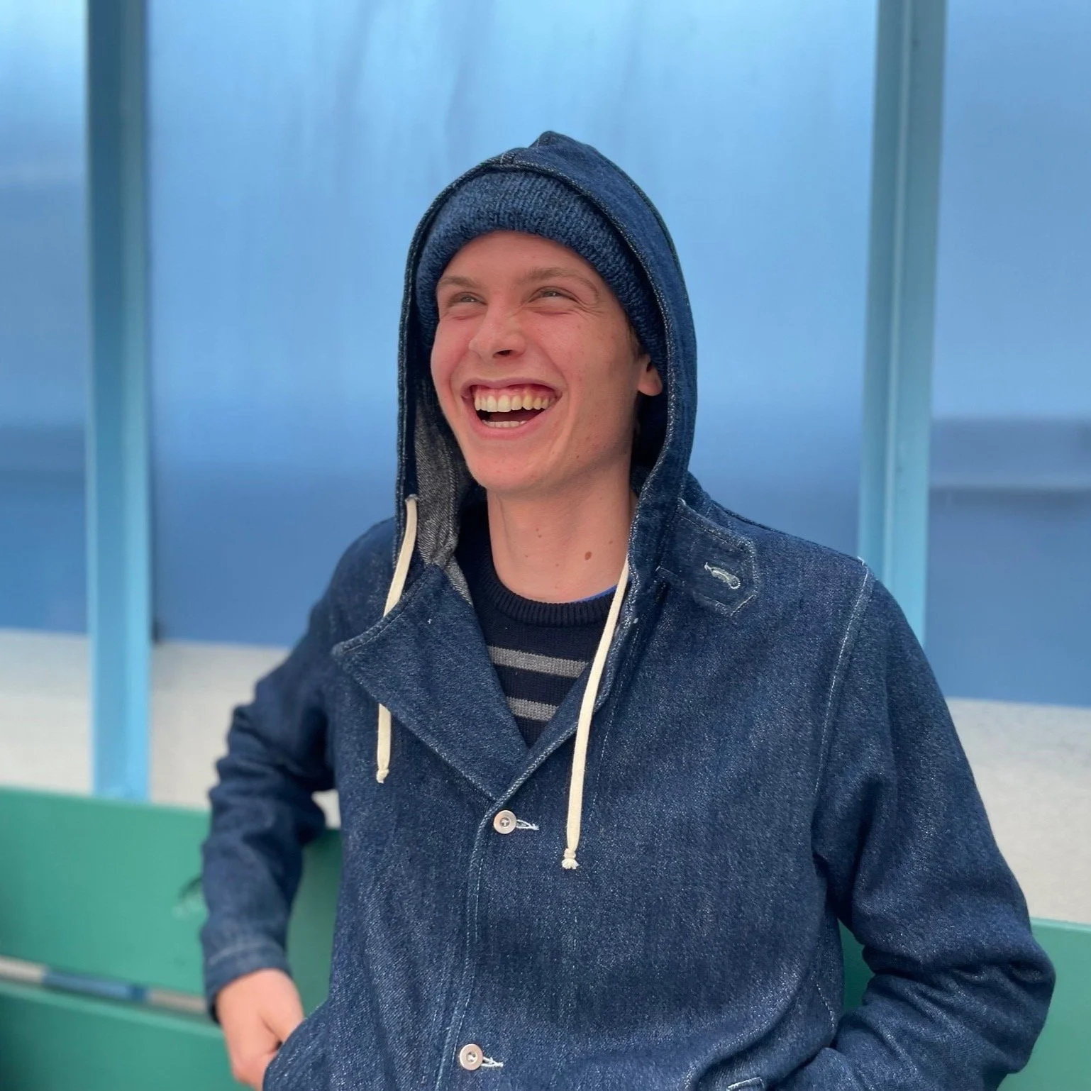 A young man with short light hair, wearing a denim jacket with the hood up and a striped shirt underneath, smiling outdoors against a blue sky and blue-paneled background.