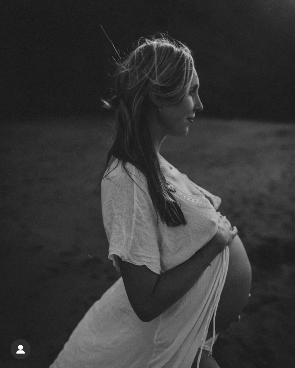 Black and white photo of a pregnant woman standing outdoors, profile view, with her eyes closed and her hand on her belly.
