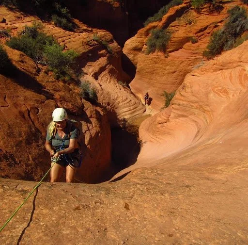 A woman wearing a white helmet and climbing gear rappelling down a rocky red canyon with lush green patches and a small group of people in the background.