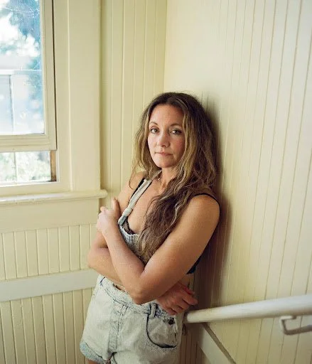A woman with long, wavy brown hair wearing a sleeveless top and high-waisted shorts, standing indoors near a window and a cream-colored wooden wall.