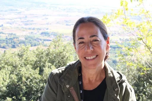 Smiling woman outdoors with trees and hills in the background.