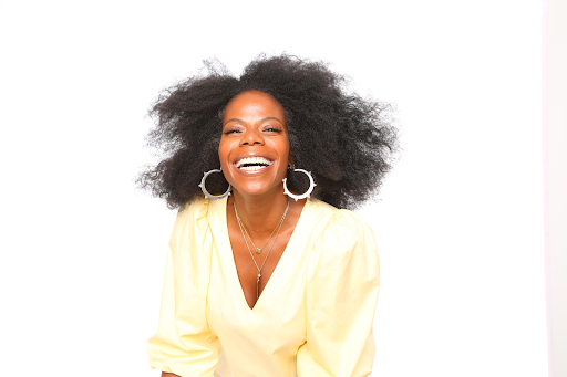 A woman with voluminous curly hair, wearing large hoop earrings, a yellow top, and a necklace, smiling brightly against a plain white background.