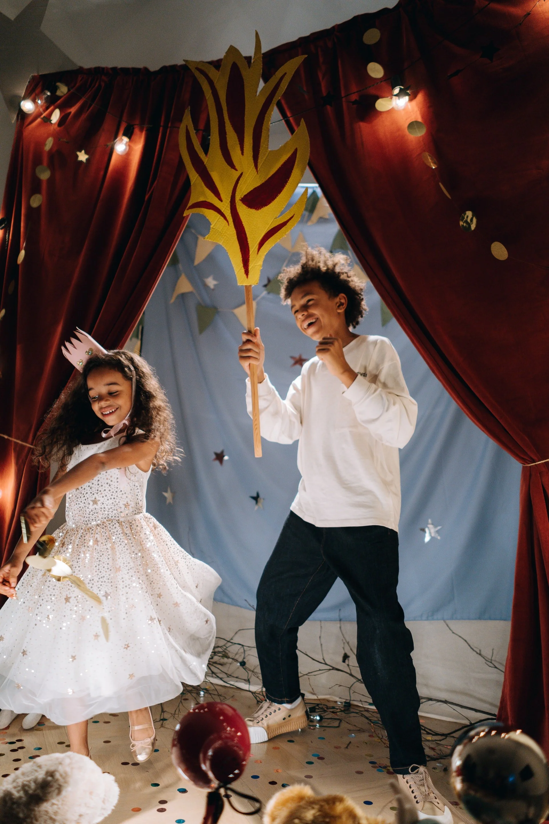 Children dressed in celebration attire playing at a party with decorations, balloons, and confetti.