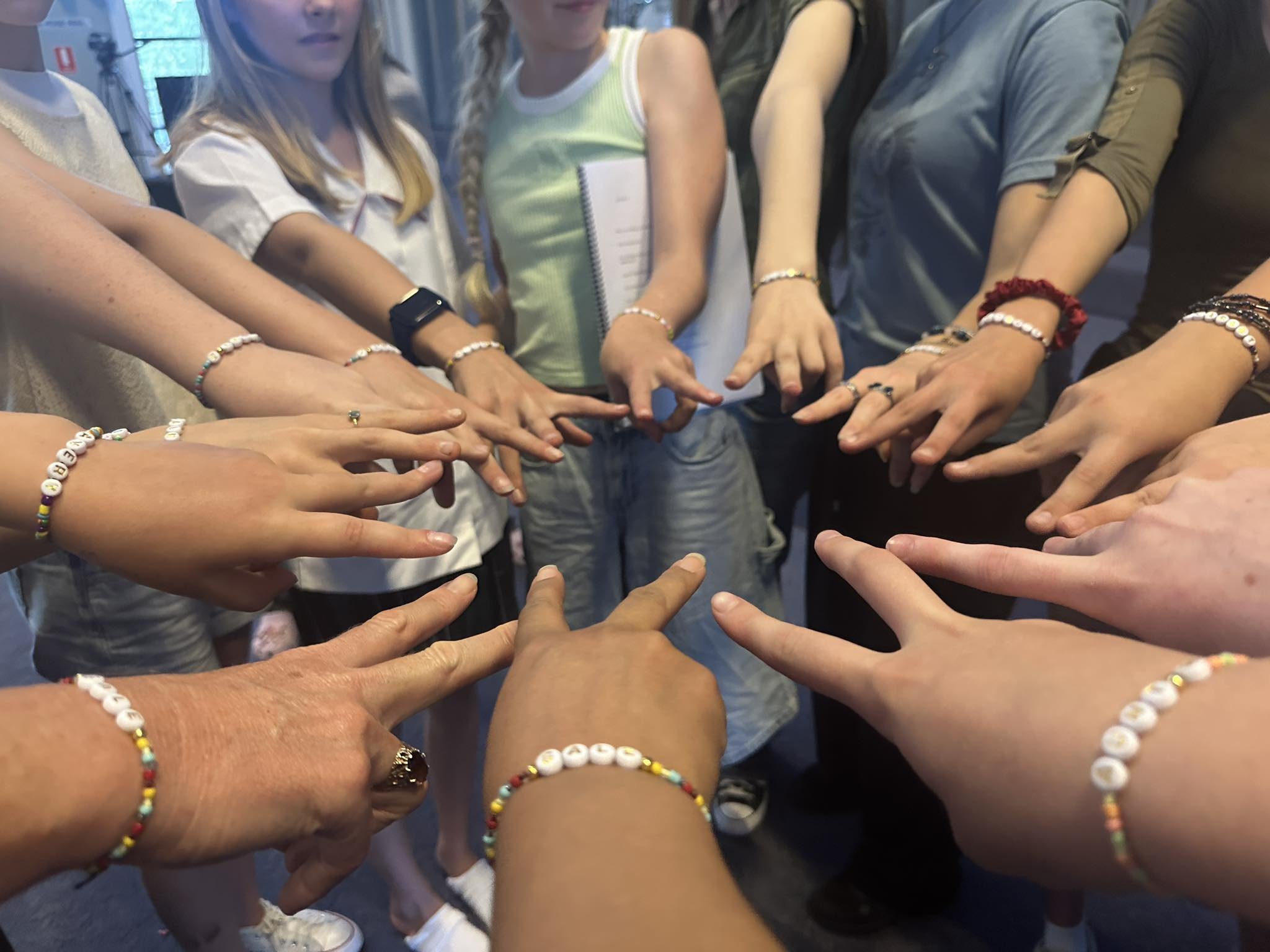 Multiple people standing in a circle with their arms extended inward, touching at the center, wearing various bracelets, in an indoor setting.