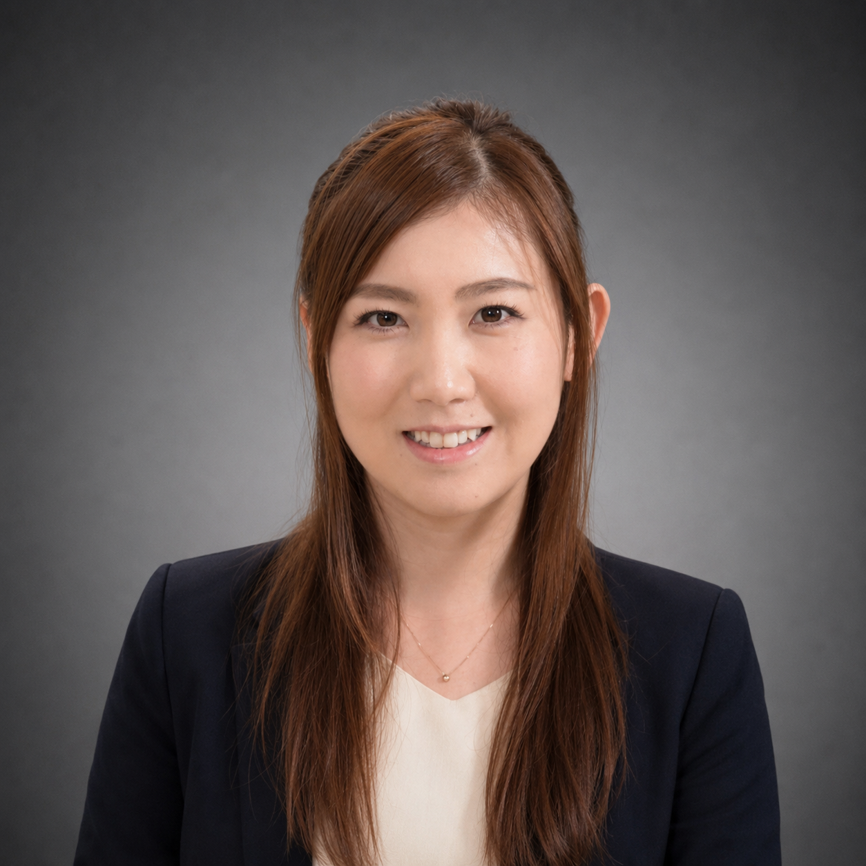 Headshot of a smiling woman with long brown hair, wearing a black blazer and white top, against a gray background.