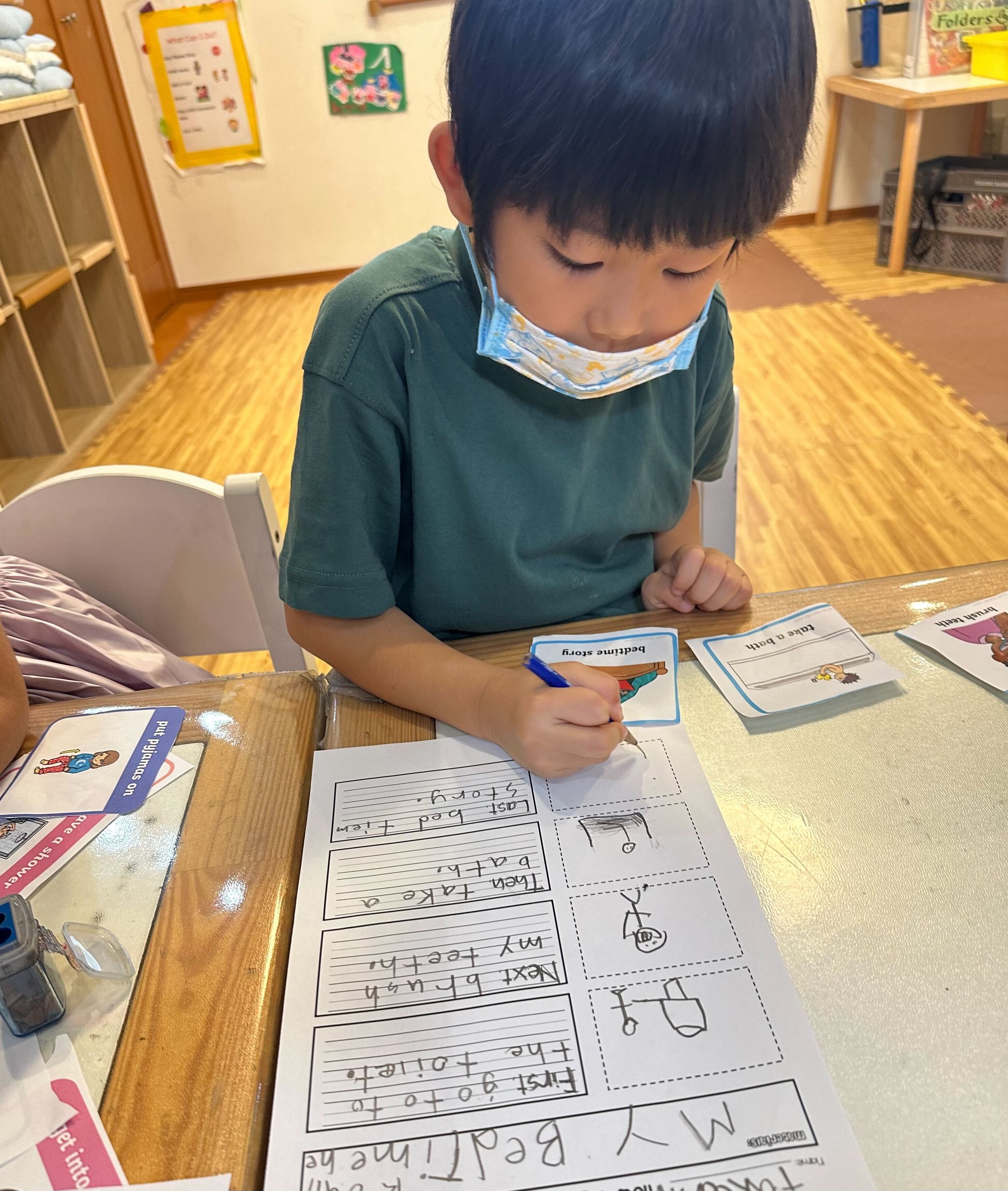 A young boy wearing a face mask, sitting at a desk, writing in a notebook. There are educational materials and picture cards on the desk.