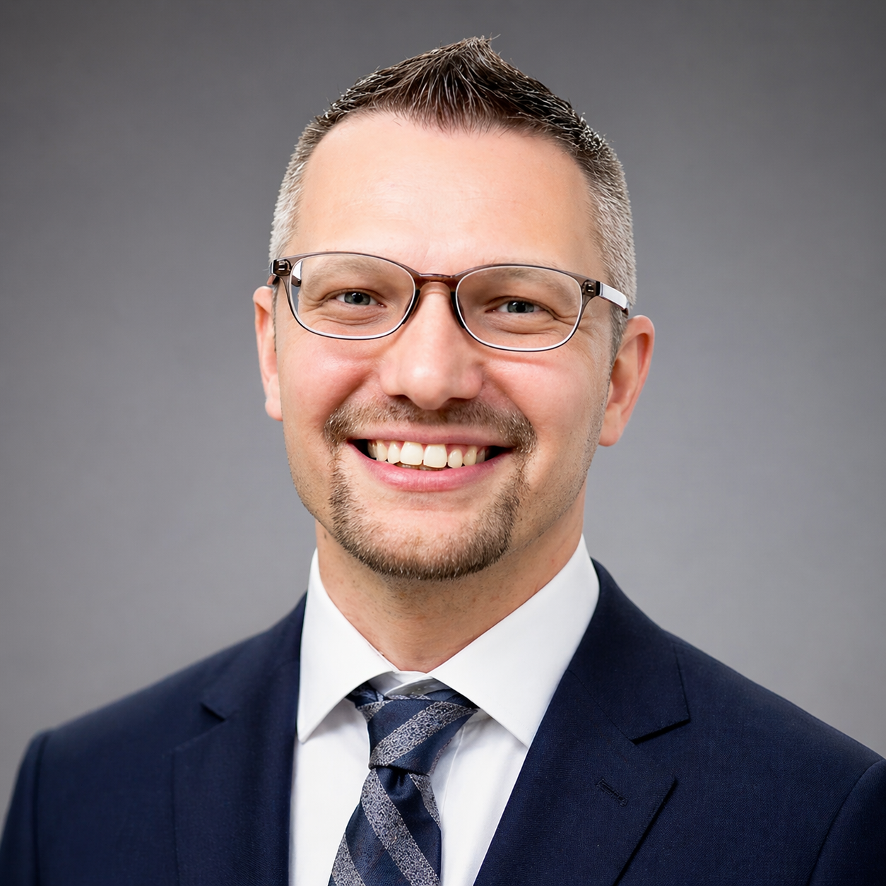 Headshot of a smiling man with glasses, facial hair, wearing a dark suit, white shirt, and a striped tie, against a plain grey background.