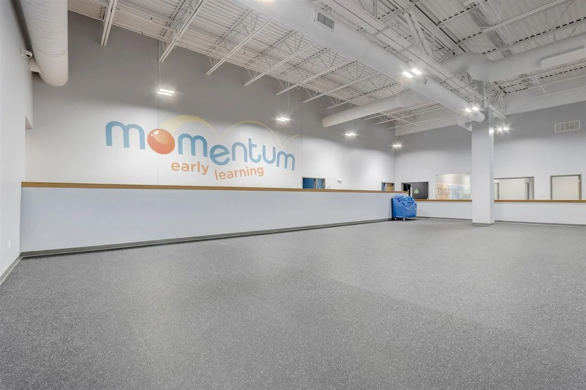 Large, empty indoor space with a wall displaying the word "momentum" with a logo and the phrase "early learning" beneath it. The room has gray carpet, white walls, an exposed ceiling with pipes and lights, and a trash bin covered with a blue tarp.