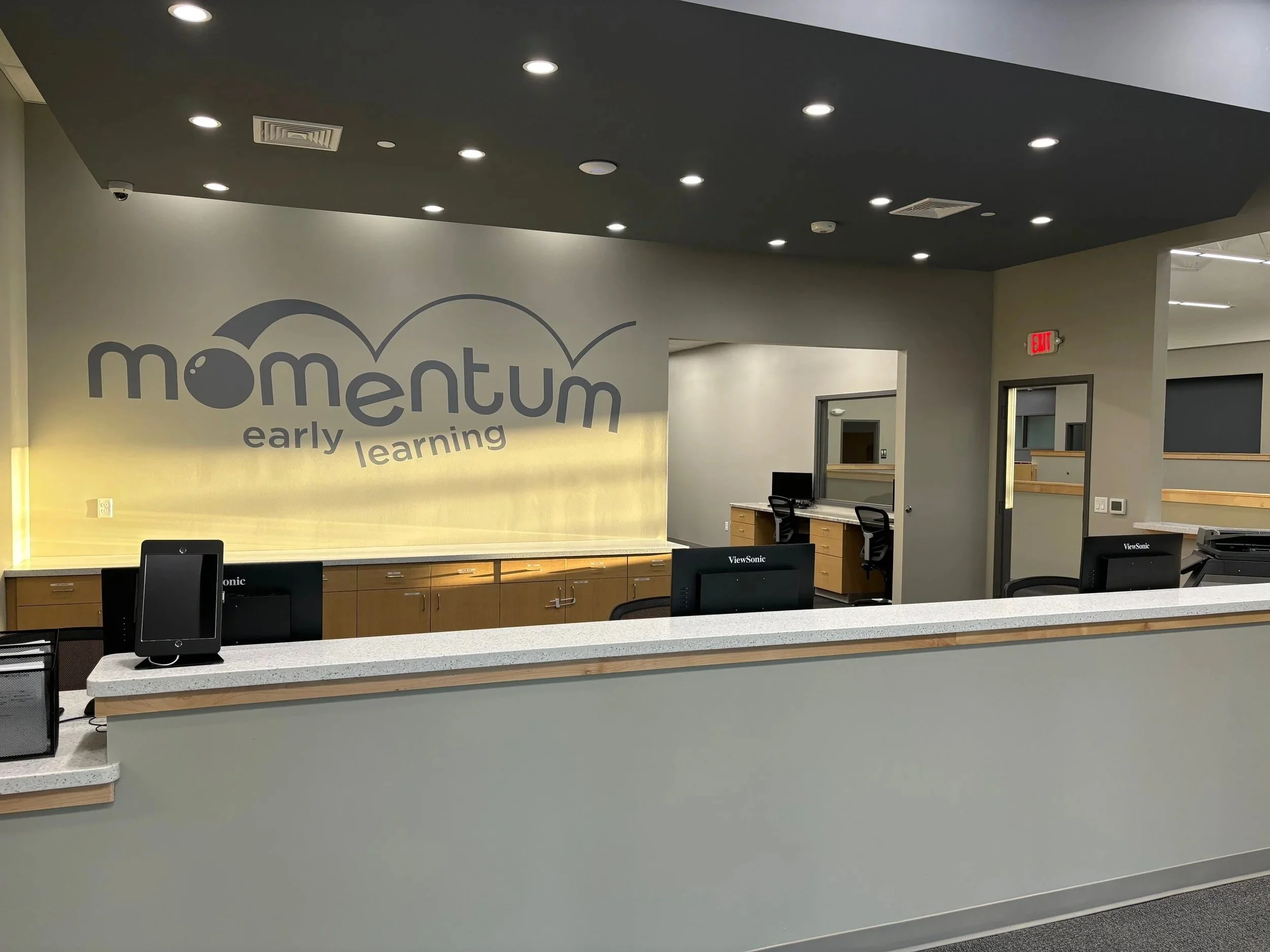 Reception area at Momentum Early Learning with a sign on the wall, computer stations, and a partial view of the hallway.