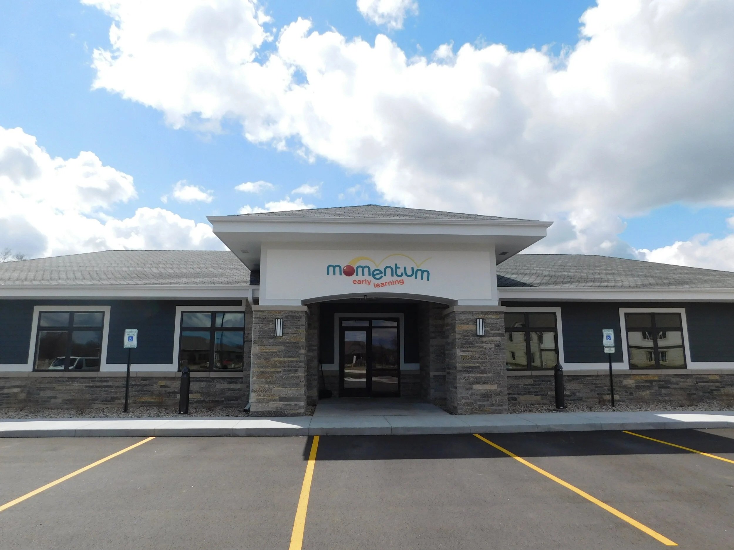 Front view of a building with a sign that reads 'Momentum early learning' above the entrance, with parking spaces in front and a partly cloudy sky overhead.