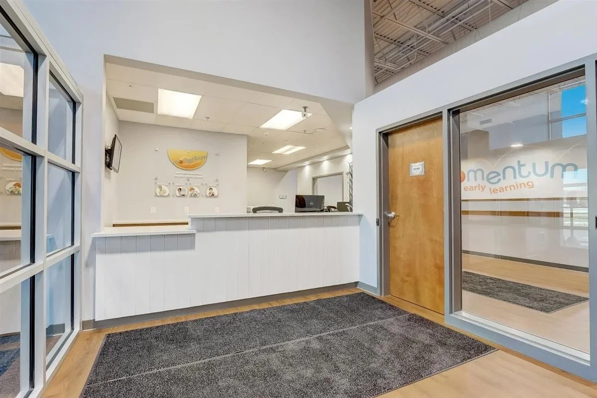 Reception area of a childhood learning center with a desk, a wall with educational posters, and glass doors showing the center's logo.