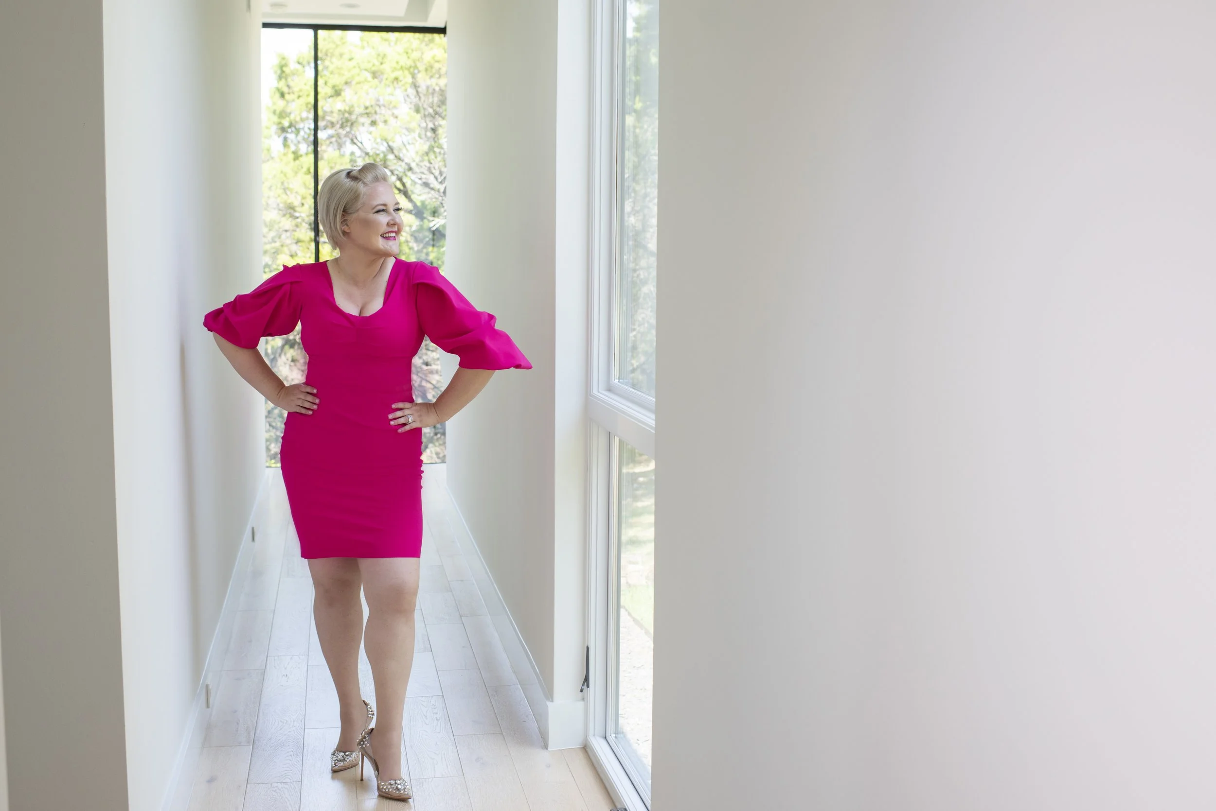 Ali Craig looking out a window in bright pink dress in sun filled hallway
