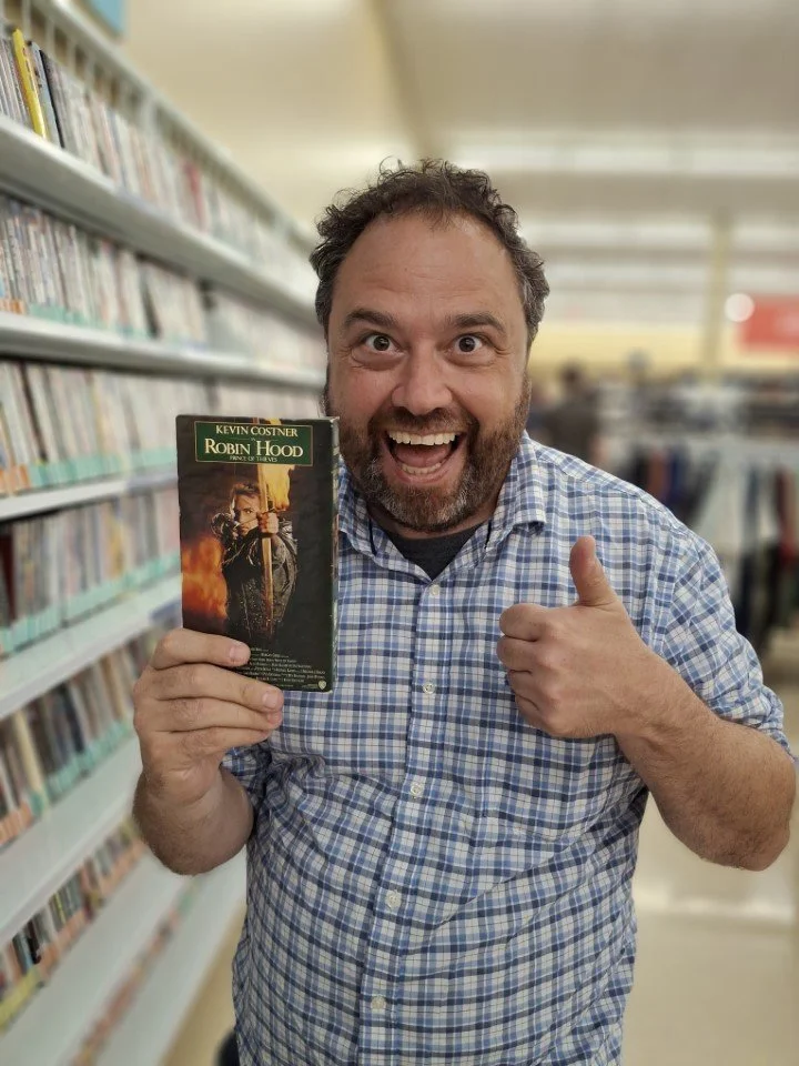 Jamie holds a “Robin Hood: Prince of Thieves” VHS at the thrift store with an excited thumbs up!