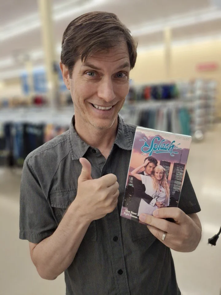 Milo holds a “Splash” movie VHS at the thrift store with a big thumbs up!