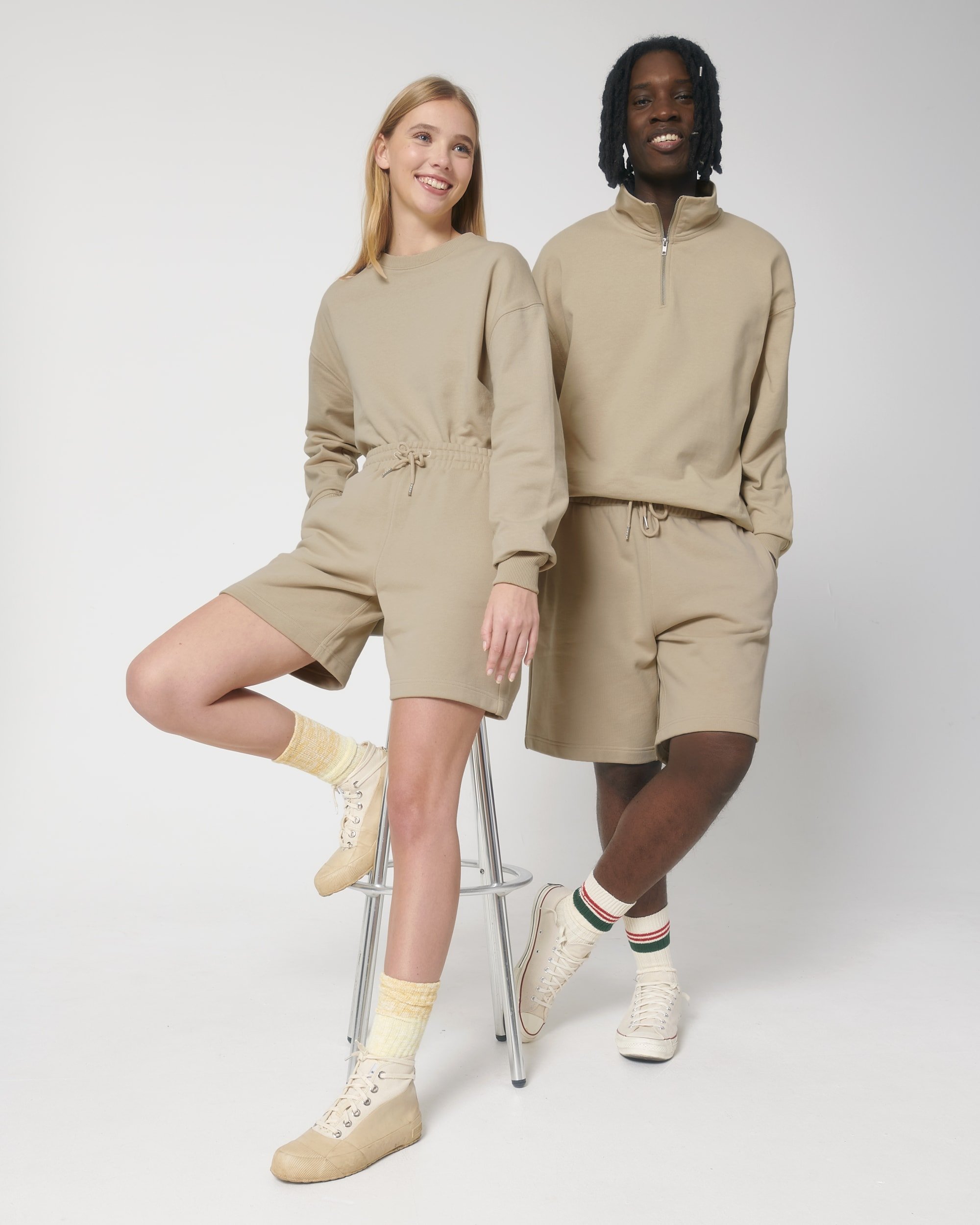 Två unga personer klädda i beige sportkläder poserar mot en vit bakgrund.