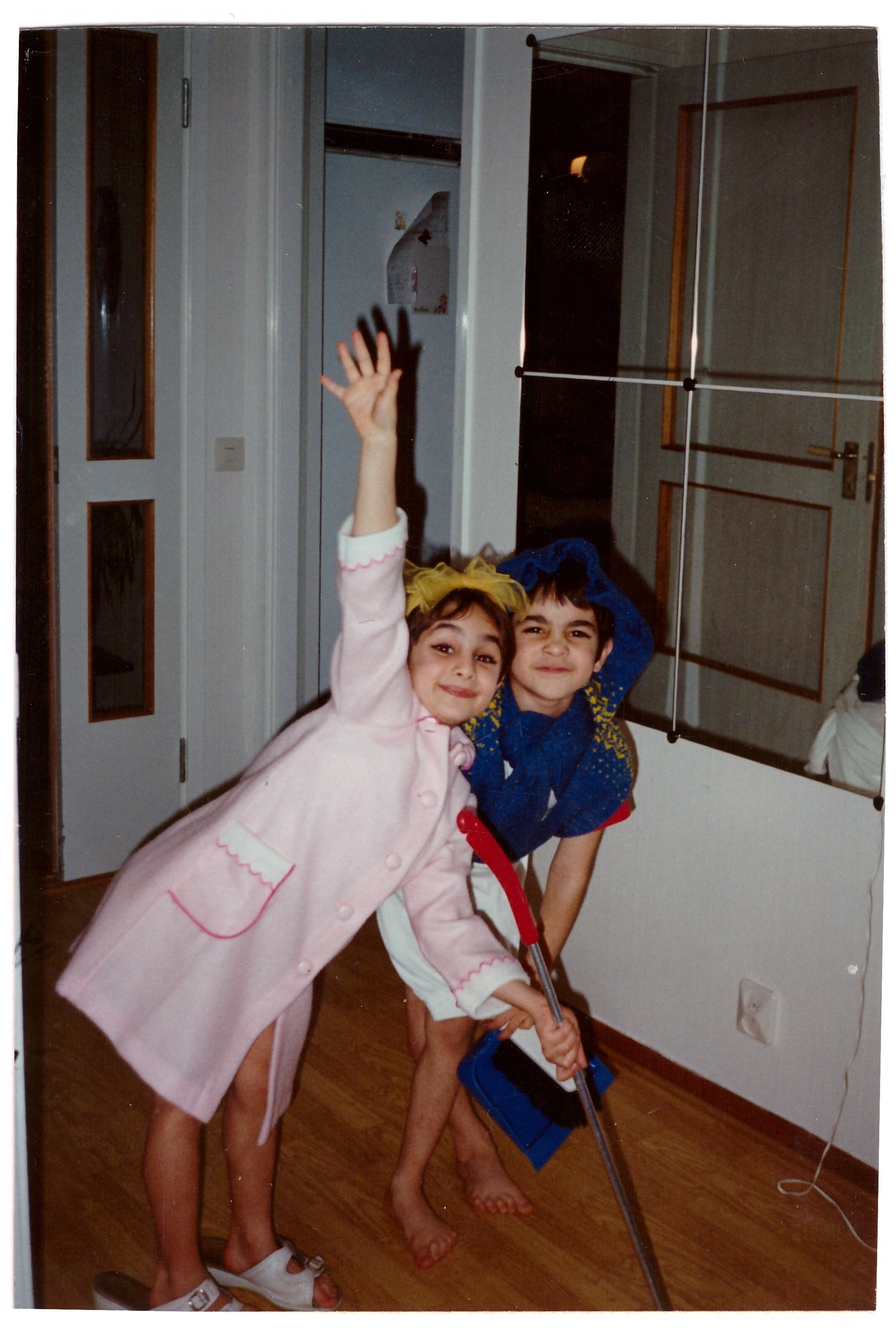 Two children playing indoors, one wearing a pink coat and white sandals, holding a dustpan, the other in a blue outfit with a yellow headband, holding a broom, smiling and posing.
