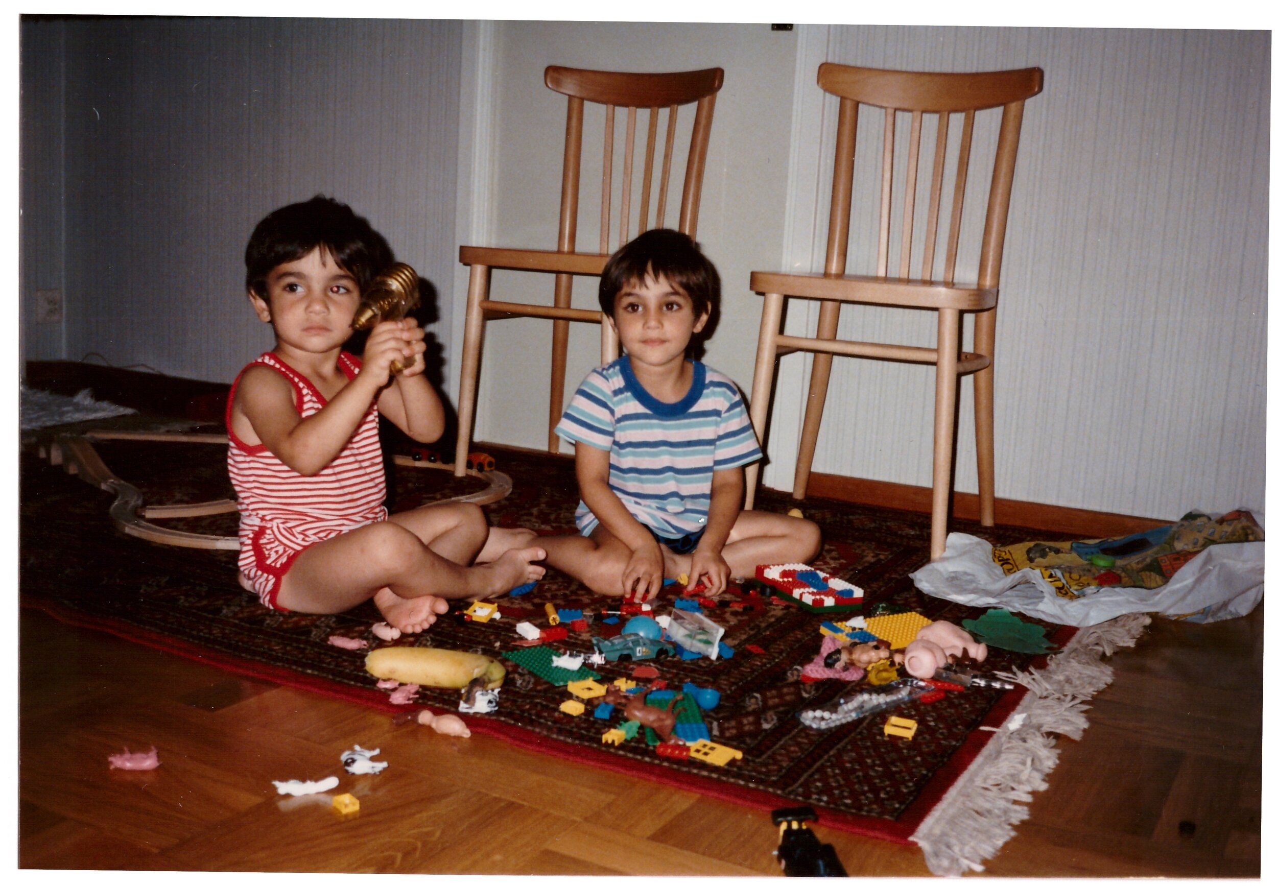 Two children sitting on a rug with toy blocks and figures, with two wooden chairs in the background.