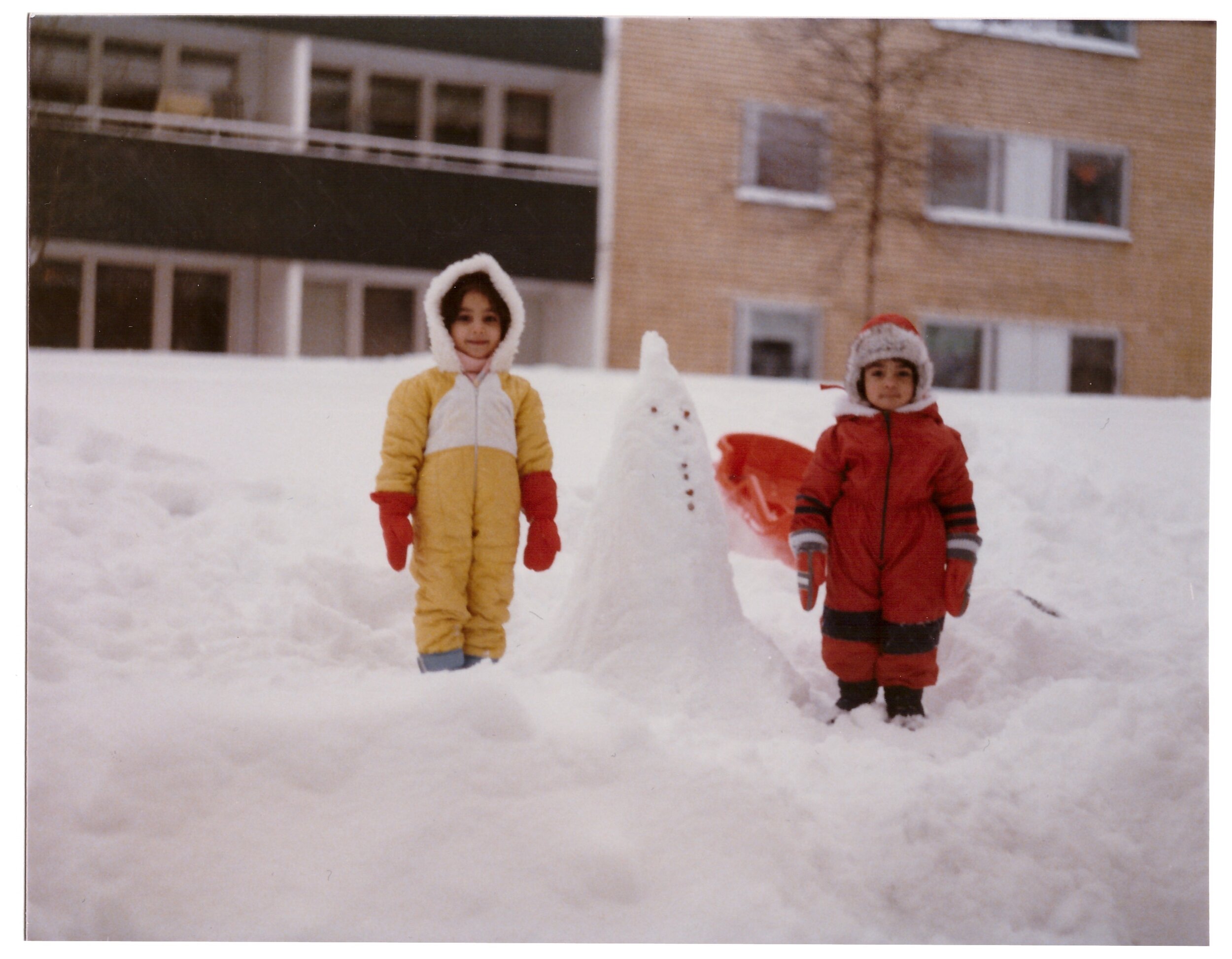 Two children in winter clothing standing in the snow beside a small snowman. The background shows an apartment building and snowy landscape.