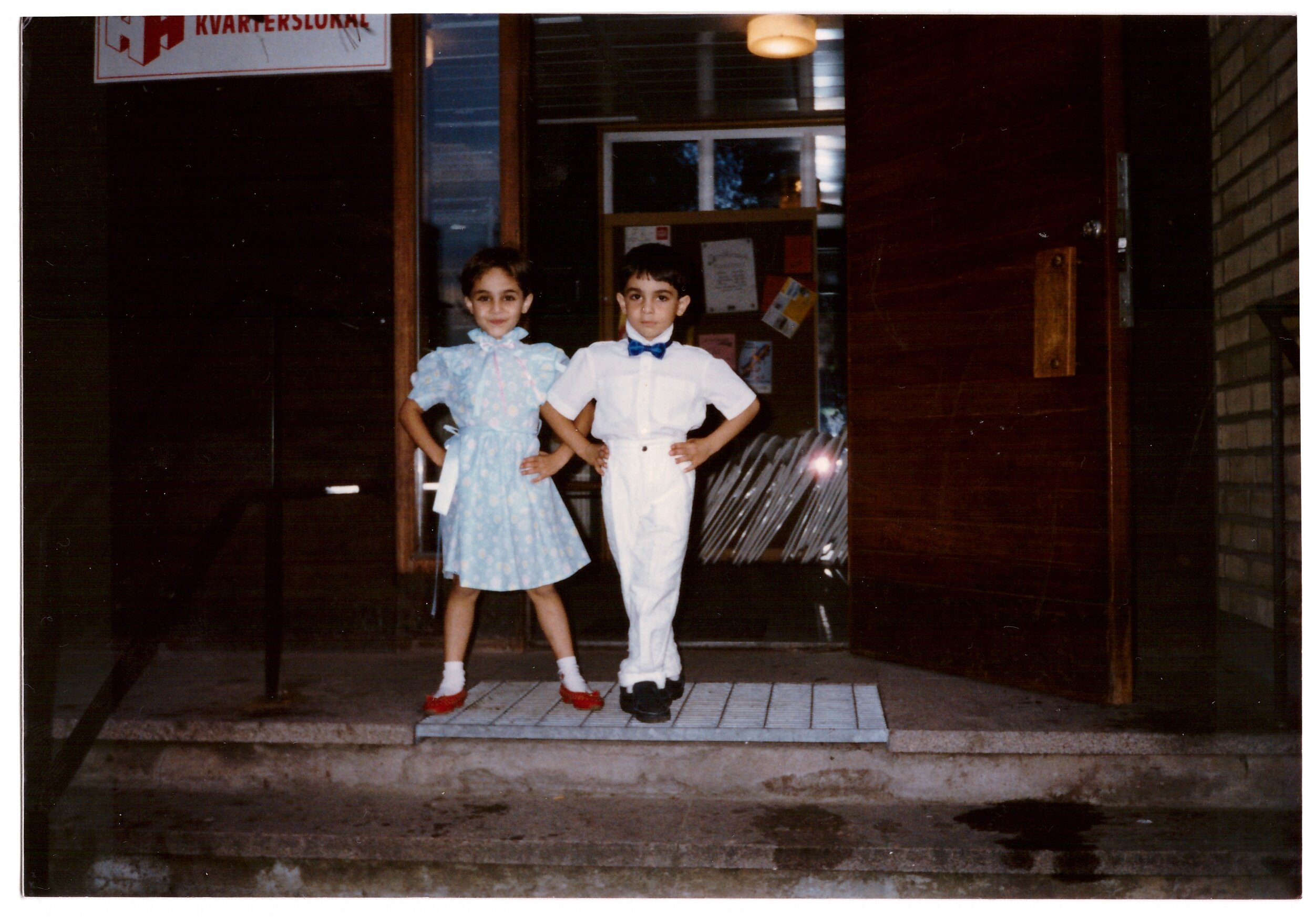 Two children posing on steps, dressed in formal attire; a girl in a light blue dress with red shoes and a boy in a white outfit with a blue bow tie.